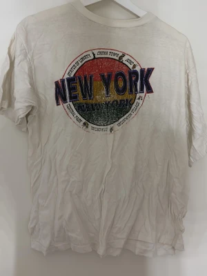 Vit New York t-shirt med tryck - Säljer en vit t-shirt med färgglatt New York-tryck på ryggen. Trycket har röda, gula och gröna toner och text med olika stadsdelar. Klassisk passform och rund hals. Perfekt för dig som gillar streetstyle och vill sticka ut. Står ej vilken storlek men den passar som en medium