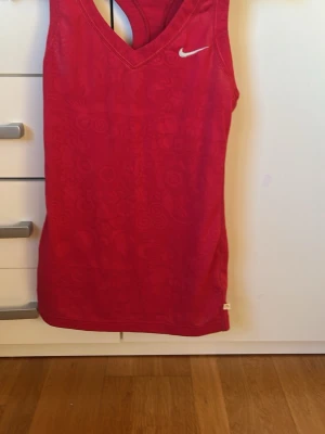 Rött sportlinne från Nike - Snyggt rött sportlinne från Nike med diskret mönster och klassisk vit logga på bröstet. Linnet har brottarrygg och är gjort i ett lätt och andningsbart material som passar perfekt för träning och sportaktiviteter.