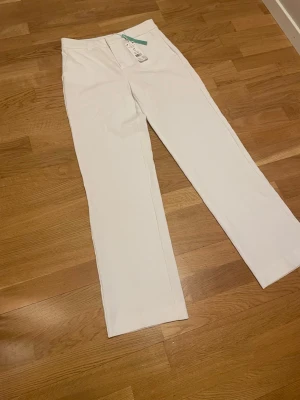 Vita raka kostymbyxor från Gina  - Säljer ett par stilrena vita kostymbyxor från gina. Byxorna har rak passform, pressveck och klassisk midja. Perfekta för en clean och fräsch look. Materialet är mjukt och har en lätt stretch för extra komfort. Taggarna sitter kvar.