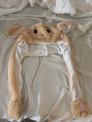 Söt kaninmössa med långa öron - Mysig och fluffig kaninmössa i beige och vitt med broderat ansikte och långa öron. Mössan har rosa tassdetaljer på ändarna och är gjord i mjuk plysch. Perfekt för dig som vill ha en gullig accessoar med djurtema.