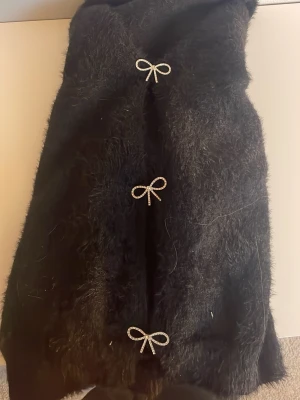 Svart pälskofta med rosettknäppning - Säljer en svart pälsjacka med lång ärm och tre dekorativa rosettknäppningar framtill. Koftan har en fluffig och mjuk yta som ger en cool look. Perfekt för dig som vill ha något unikt och stilrent i garderoben.