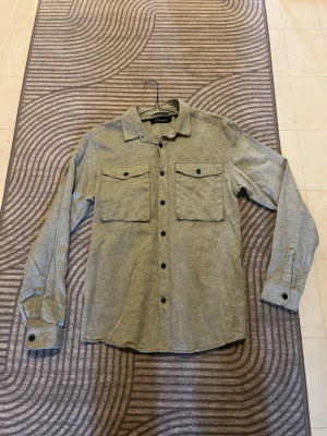 Ljusgrå skjorta/overshirt från peakperformance - Snygg ljusgrå skjorta med två stora bröstfickor och svarta knappar hela vägen fram. Skjortan har klassisk krage och långa ärmar med knapp vid manschetten. Perfekt för en avslappnad men stilren look. Passar även snyggt som en overshirt🙌