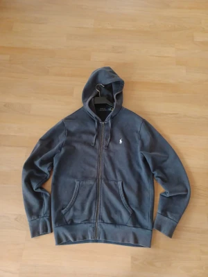 Mörkgrå hoodie från Polo Ralph Lauren - Mörkgrå hoodie med dragkedja från Polo Ralph Lauren. Klassisk design med två fickor fram, huva med snören och broderad logotyp på bröstet. Materialet är mjukt och bekvämt, perfekt för en avslappnad stil.