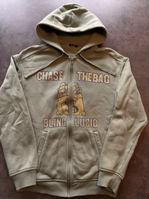 Grön hoodie med bling och tryck - Stilren ljusgrön hoodie med dragkedja och huva från Bling Lucid. Framsidan har blingade stenar som bildar texten 'CHASE THE BAG' och 'BLING LUCID' samt en stor pengasäck i guld och svart. Perfekt för dig som gillar streetwear och detaljer.