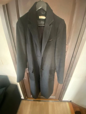 Svart klassisk ullkappa - Stilren svart ullkappa med två knappar framtill, breda slag och fickor på sidorna. Lång modell som ger en elegant siluett. Perfekt för dig som vill ha en tidlös och snygg look till höst och vinter.