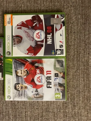 Xbox 360 spel FIFA 11 & NHL 09 - Två Xbox 360-spel: FIFA 11 med fotbollstema och NHL 09 med ishockeytema. Båda spelen har gröna plastfodral och tydliga EA Sports-loggor på framsidan. FIFA 11 har en röd och vit design, NHL 09 har röd och vit hockeytröja på omslaget.
