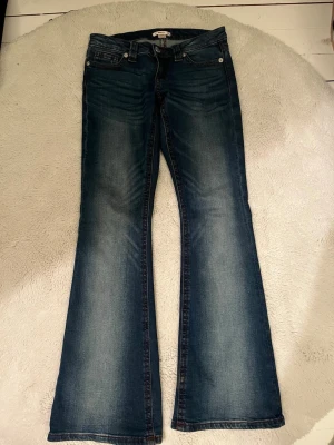 Blå bootcut jeans med röd söm  - Säljer ett par blå bootcut jeans  med klassiska kontrastsömmar och broderade bakfickor. Jeansen har en snygg tvätt med slitningar framtill och är tillverkade i denim med lätt stretch. Perfekta för dig som gillar retrovibbar och markerad siluett.