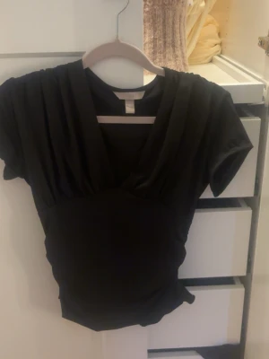 Svart omlottopp från H&M - Snygg svart omlottopp från H&M med korta ärmar och djup v-ringning. Toppen har en figurnära passform och är tillverkad i mjukt, stretchigt material som sitter bekvämt. Perfekt att styla med jeans eller kjol för en trendig look.