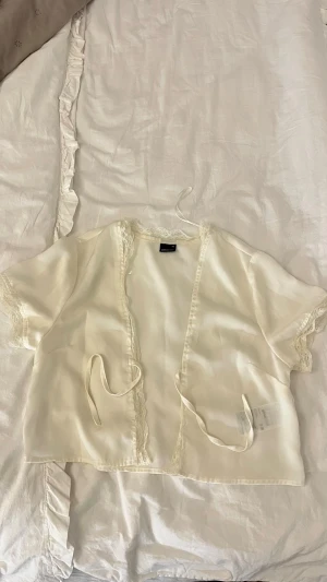 Krämvit kimono från Gina Tricot - Söt krämvit kimono i satin från Gina Tricot med korta ärmar och spetsdetaljer längs kanterna. Knytband framtill och mjukt, glansigt material som känns lyxigt mot huden. Perfekt för chill dagar hemma eller som loungewear.