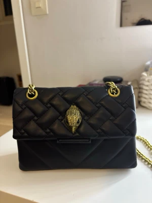 KURT GEIGER VÄSKA - Stilren svart quiltad handväska med guldfärgade kedjor och detaljer. Väskan har ett dekorativt emblem framtill och magnetlås. Perfekt för dig som gillar klassisk men trendig design. Kedjan gör att den kan bäras både på axeln och crossbody.