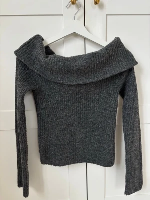 Mörkgrå off shoulder stickad tröja - Mysig mörkgrå ribbstickad tröja med bred off shoulder-krage och långa ärmar. Tröjan har en kroppsnära passform och är perfekt för dig som gillar en stilren och trendig look. Materialet känns mjukt och värmande, perfekt för kyligare dagar.