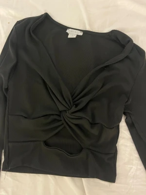 Svart twistad croppad topp MISU - Trendig svart croppad topp från MISU med twistad detalj framtill och v-ringad hals. Toppen är ribbad och har långa ärmar samt en liten cut-out under tvisten. Perfekt för dig som gillar en edgy och modern stil.