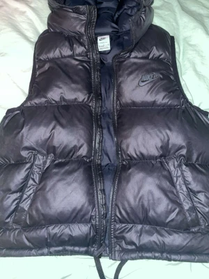 Svart dunväst från Nike - Snygg svart dunväst från Nike med hög krage och dragkedja framtill. Västen har två snedställda fickor och diskret Nike-logga på bröstet. Tillverkad i glansigt material och fodrad för extra värme. Perfekt för lager-på-lager och streetwear-stil.