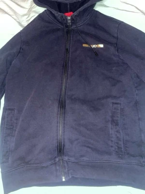 Svart zip up med dragkedja från Hugo Boss - Säljer en svart hoodie från Hugo Boss med dragkedja framtill och två fickor. Hoodien har huva med snören och ett diskret Hugo-emblem på bröstet. Perfekt för dig som gillar stilrena och exklusiva plagg. Materialet är mjukt och bekvämt.