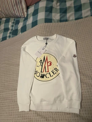 Vit sweatshirt från Moncler - Snygg vit sweatshirt från Moncler med stort tryck på bröstet i beige, svart och rött. Tröjan har rund hals, långa ärmar och ribbade muddar. Moncler-logga på ärmen ger en exklusiv touch. Perfekt för dig som gillar streetwear och premiumkänsla.