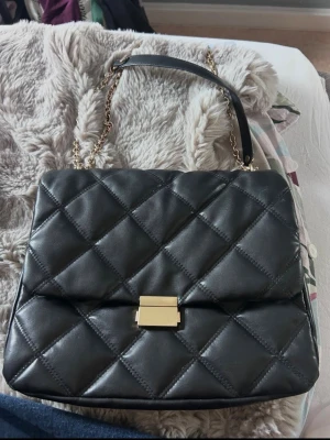 Svart quiltad handväska med kedja - Snygg svart handväska i quiltat mönster med guldfärgat spänne och axelrem i kedja och läder. Klassisk rektangulär form och elegant design som passar till många olika stilar. Perfekt accessoar för dig som gillar trendiga detaljer.