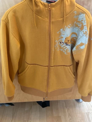Quiksilver hoodie  - Säljer en gul hoodie med hel dragkedja och coolt vitt tryck på bröstet. Hoodien har två fickor fram, ribbade muddar och en stor huva med mjukt foder. Perfekt för dig som vill ha en skön och snygg hoodie till vardags.