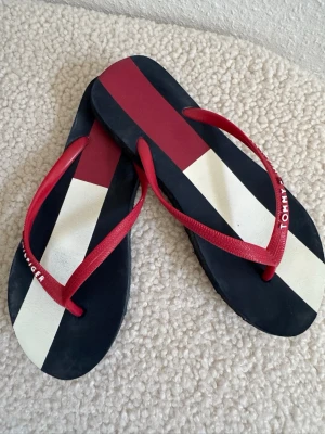 Randiga flip flops från Tommy som nya Hilfiger - Snygga flip flops från Tommy Hilfiger med breda ränder i marinblått, vitt och rött. Remmarna är röda och sandalerna har en klassisk, stilren design. Perfekta för sommaren och strandhäng. Tillverkade i syntetmaterial. Storlek 7-8 amerikansk som motsvara 37,5-38,5. Innelängd 24,5cm.
