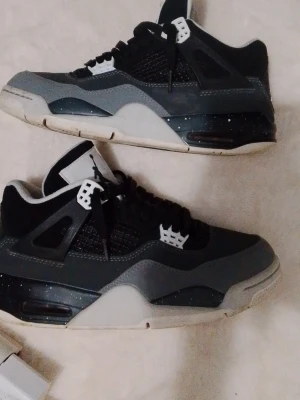 Svarta och grå Air Jordan 4 sneakers - Säljer ett par Air Jordan 4 sneakers i svart och grått med vita detaljer och svart mesh. Skorna har den klassiska höga siluetten, synlig Air-enhet i sulan och Jumpman-logga på plösen. Materialet är en mix av mocka och textil med plastdetaljer.