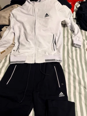 Adidas svartvitt träningsset - Snyggt svartvitt träningsset från Adidas med zip-jacka och matchande byxor. Jackan har dragkedja, ståkrage och svarta detaljer vid muddar och fickor. Byxorna har vita dragkedjor och Adidas-logga på benet och bakfickan. Perfekt för träning eller chill. Köpt från Turkiet, dvs inte äkta därav priset!