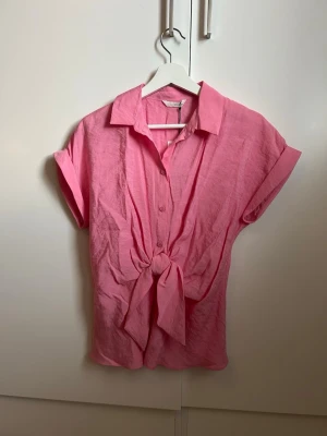 Rosa knytblus från Zara - Snygg rosa blus från Zara med korta ärmar och knytning framtill. Blusen har klassisk krage, knappar hela vägen och är gjord i ett lätt, luftigt material som passar perfekt till sommaren. Enkel men trendig design som lyfter vilken outfit som helst.
