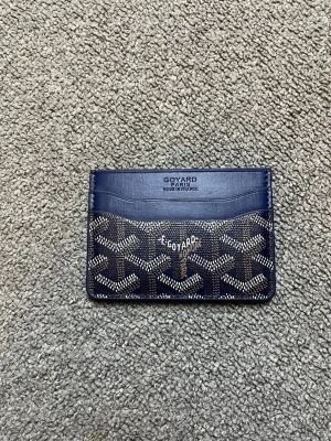 Mörkblå kortfodral från Goyard - Snyggt mörkblått kortfodral från Goyard med klassiskt mönster i vitt och brunt. Fodralet har flera kortfack både fram och bak och är tillverkat i exklusivt läder med canvasdetaljer. Perfekt för dig som vill ha en stilren och lyxig accessoar.