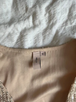 Glittrig beige topp från Nelly - Säljer en glittrig beige topp från Nelly med långa ärmar och knyt i midjan. Toppen har paljetter över hela och en croppad passform som ger en trendig look. Perfekt för dig som vill sticka ut lite extra.