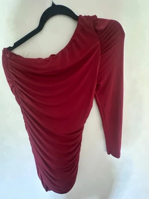 Röd oneshoulder klänning  - Snygg röd oneshoulder klänning från WAL G med draperad effekt och tight passform. Klänningen har en lång ärm och är tillverkad i ett mjukt, stretchigt material som framhäver figuren. Perfekt för dig som vill sticka ut med en unik och trendig look.