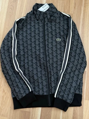Svart mönstrad track jacket Lacoste - Säljer en svart track jacket från Lacoste med grått geometriskt mönster och vita ränder längs ärmarna. Dragkedja framtill, ribbade muddar och klassisk Lacoste-logga på bröstet. Perfekt för en sportig och trendig look.