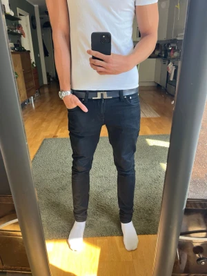 Nudie jeans - Säljer ett par svarta slim fit jeans med klassisk femficksdesign och smal passform. Jeansen har normal midja och är tillverkade i ett stretchigt material som sitter snyggt på benen. Perfekta för dig som gillar en stilren och modern look.