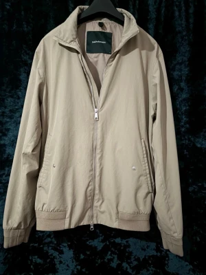 Beige peak performance vindjacka med dragkedja - Superfin och stilren beige jacka från Peak Performance i bombertillskärning med ribbade muddar. Tillverkad i Peaks högkvalitativa HIPE-material vilket gör den 100% vindtät, vattentät (5 000 mm) samt med god andningsförmåga. Perfekt jacka för både vår, sommar och höst! Jackan har en snygg mattsvart gummiloga på ärmen, två sidofickor med tryckknappar samt en praktisk innerficka med dragkedja.  Märke: Peak Performance  Färg: Beige / Sand  Storlek: M (passar S)  Skick: Mycket fint skick!