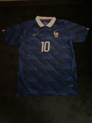 Blå Frankrike fotbollströja Nike - Säljer en blå Frankrike fotbollströja från Nike med nummer 10 på bröstet. Tröjan har vita detaljer, klassisk krage och är tillverkad i Dri-FIT-material som andas. Perfekt för fotbollsträning eller att bära som supporter. Snyggt mönster och officiell logga på bröstet.
