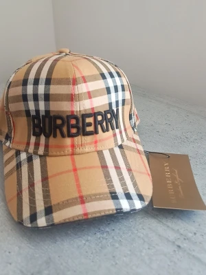 Burberry beige keps perfekt för sommaren  - Snygg keps från Burberry i klassiskt beige rutigt mönster med svart broderad logga framtill och broderad ryttare bak. Justerbar rem bak för perfekt passform. Tillverkad i bomull för en bekväm känsla och stilren look.