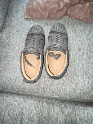 Svarta sneakers med nitar och röd sula - Unika svarta sneakers med spetsiga nitar över tån och cool röd sula. Skorna har snörning och är tillverkade i ett material som liknar läder. Insidan är beige och designen är riktigt edgy och sticker ut i mängden. har använt dom under vintern men kommer inte använda dom något mer. PRIS KAN DISKUTERAS!!!