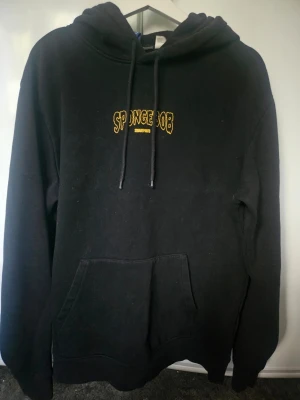Svart Spongebob hoodie med tryck - Svart hoodie med huva och dragsnören, Spongebob-tryck i gult framtill och stort färgglatt motiv på ryggen med texten 'I TOUCHED MY BRAIN'. Känguruficka framtill och avslappnad passform. Perfekt för dig som gillar streetwear och tecknade serier.