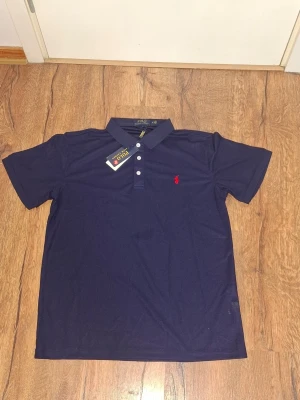 Mörkblå pikétröja från Polo Ralph Lauren - Klassisk mörkblå pikétröja från Polo Ralph Lauren med röd broderad logga på bröstet. Tröjan har korta ärmar, krage och knappar framtill. Perfekt för en stilren och avslappnad look.