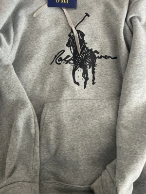 Grå hoodie från Polo Ralph Lauren - Snygg rep grå hoodie från Polo Ralph Lauren med stor svart logga och polospelare på bröstet. Klassisk känguruficka framtill och justerbar huva med gråa snören. hoodien är tillverkad i mjuk material 