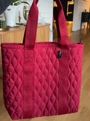 Röd quiltad shoppingväska Victoria's Secret - Säljer en oanvänd snygg röd quiltad shoppingväska från Victoria's Secret med breda axelband där loggan är invävd. Väskan har ett mjukt material och rymlig form, perfekt för att bära allt du behöver, med två innerfickor. Stängs med dragkedja och har en modern, trendig look. Ursprungspris: ca 500kr💞