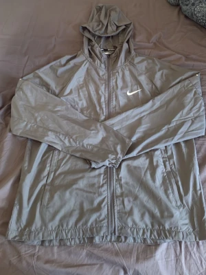 Grå vindjacka med huva från Nike - Snygg grå vindjacka från Nike med huva och dragkedja framtill. Jackan har två sidofickor med dragkedja och en vit Nike-logga på bröstet. Tillverkad i lätt och vindtätt material, perfekt för blåsiga dagar.
