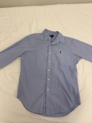 Blårandig skjorta från Polo Ralph Lauren - Klassisk blårandig skjorta från Polo Ralph Lauren med button-down krage och lång ärm. Skjortan har vita knappar framtill och den ikoniska broderade loggan på bröstet. Tillverkad i mjuk bomull för en bekväm känsla.