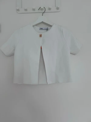 Vit kortärmad cardigan från Zara - Stilren vit cardiganjacka från Zara med korta ärmar och rund halsringning. Jackan har en enkel knapp framtill och rak passform. Perfekt för en clean och modern look.