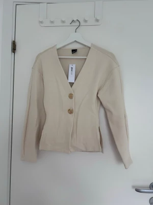 Beige kofta med guldknappar - Stilren beige kofta med djup V-ringning och två dekorativa guldknappar framtill. Koftan har långa ärmar och rak passform, perfekt för en enkel men trendig look. Tillverkad i ett mjukt material som känns skönt mot huden.