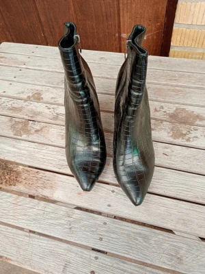 Svarta croco-mönstrade stövletter stl 42 - Säljer ett par svarta stövletter med croco-mönster och spetsig tå. Skorna har dragkedja på insidan och en smal siluett som ger en edgy look. Materialet ser ut som glansigt konstläder med struktur. Perfekta för dig som vill sticka ut med dina boots.