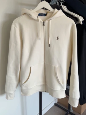 Krämvitt set från Polo Ralph Lauren - Snyggt krämvitt set från Polo Ralph Lauren med hoodie och matchande byxor. Hoodien har dragkedja, fickor och logga på bröstet. Byxorna har resår i midjan och muddar vid benslut. Perfekt för en avslappnad och stilren look.