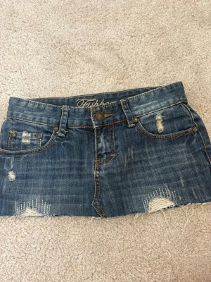 Blå jeanskjol från Fishbone - Säljer en kort jeanskjol från Fishbone i blå denim med slitna detaljer och rå kant nertill. Kjolen har klassiska fem fickor, bälteshällor och en knapp framtill. Perfekt för dig som gillar en avslappnad och trendig stil.💓