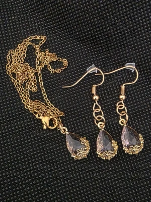 Set med guldfärgat halsband och örhängen - Säljer ett matchande smyckeset med guldfärgat halsband och örhängen. Både halsbandet och örhängena har droppformade, ljusrosa stenar infattade i detaljerade guldfärgade metallhylsor med små blommor. Perfekt för dig som gillar elegant och glittrig stil.