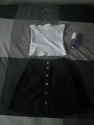 Svart kortkjol med knappar framtill - Snygg svart kortkjol med raka linjer och silverfärgade knappar längs hela framsidan. Kjolen har en enkel och stilren design som passar till många olika outfits. Materialet känns som bomull eller polyester och modellen är normal i passformen.