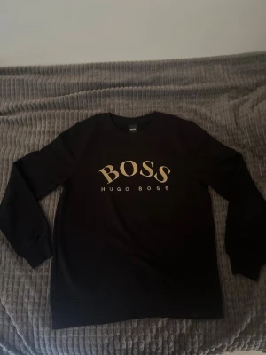 Svart sweatshirt från Hugo Boss - Svart sweatshirt från Hugo Boss med stort BOSS-tryck i vitt och guld på bröstet. Klassisk rund halsringning och långa ärmar. Perfekt för en avslappnad och stilren look.