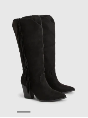 Svarta cowboy boots med fransar - Snygga svarta cowboy boots i mockaimitation med högt skaft och fransdetaljer längs sidan. Dessa är nya och inte använda!