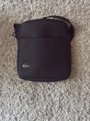 Svart axelväska från Lacoste - Snygg svart axelväska från Lacoste med klassisk krokodillogo i silver på framsidan. Väskan har en fyrkantig form, justerbar axelrem och är tillverkad i slitstarkt tygmaterial. Perfekt för att bära det viktigaste med stil.